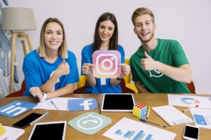 Young digital marketing team promoting social media platforms with Instagram icon, thumbs up gestures, and branding materials on the table.